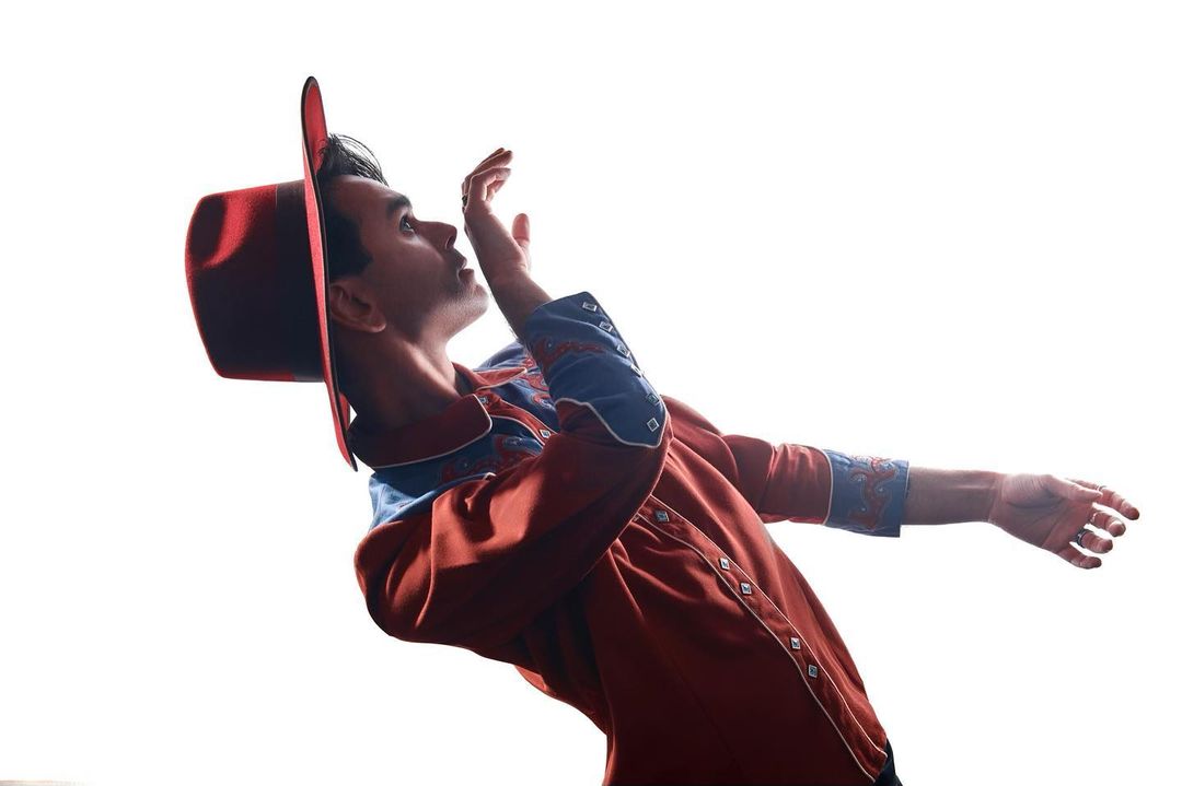 Man in a red hat and rust-colored western shirt leans back with one arm raised against a pure white background in a studio shot.